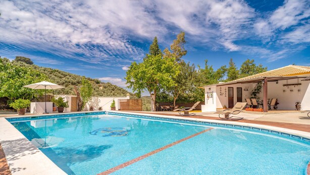 Une piscine à l’eau d’un bleu cristallin, entourée de chaises longues et de parasols, avec vue sur une colline et un bâtiment doté d’un toit en tuiles.