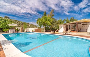 A swimming pool with clear blue water, surrounded by lounge chairs and umbrellas, with a view of a hillside and a building with a tiled roof. - Pet friendly home in Iznajar (Iznajar)