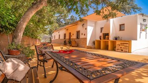 Une terrasse avec une table et des bancs en bois, entourée de plantes en pot et d’un bâtiment blanc doté de fenêtres et de portes.