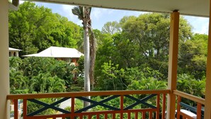 Une vue depuis un balcon doté d’une balustrade en bois, donnant sur un jardin luxuriant avec différents arbres et un bâtiment blanc en arrière-plan.