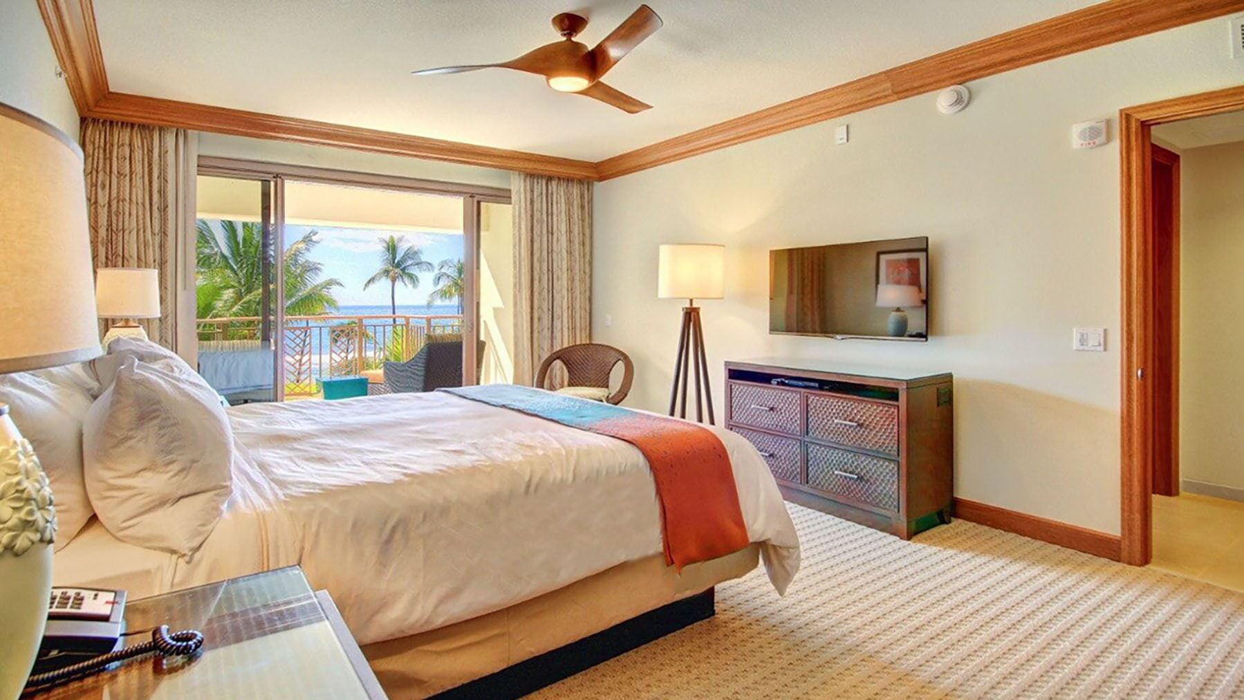 Habitación de hotel con cama, escritorio con teléfono, televisor y balcón con vista al océano.
