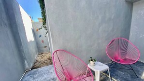 Dues cadires amb estructura de filferro rosa, un taula petita blanca i una planta en test, tot en un espai exterior reduït amb parets grises i un terra de formigó.
