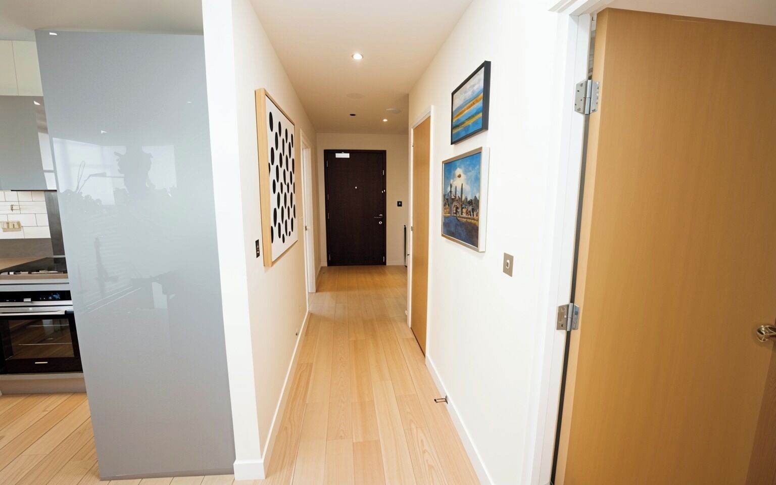 A hallway with wooden flooring, a kitchen area on the left, and a door at the end.