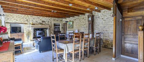 A rustic interior with a stone wall, wooden ceiling beams, a dining table with chairs, and a fireplace.