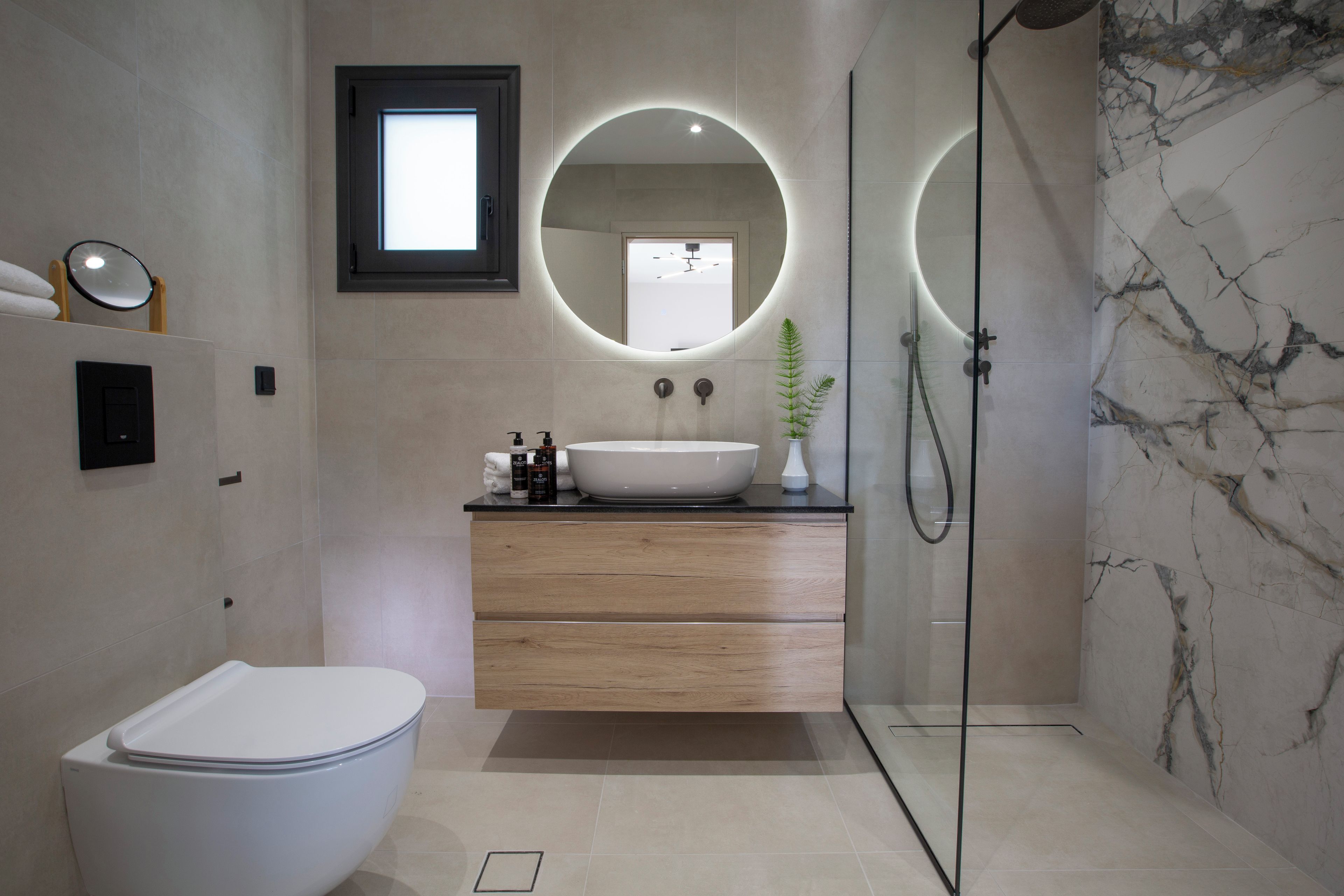 Een moderne badkamer met een wit toilet, een houten wastafelmeubel en een grote spiegel met ronde verlichting.