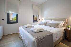A bedroom with a bed, two windows with blinds, a bedside table with a plant, and a light fixture. - Pomaria Residences (Lefkada)