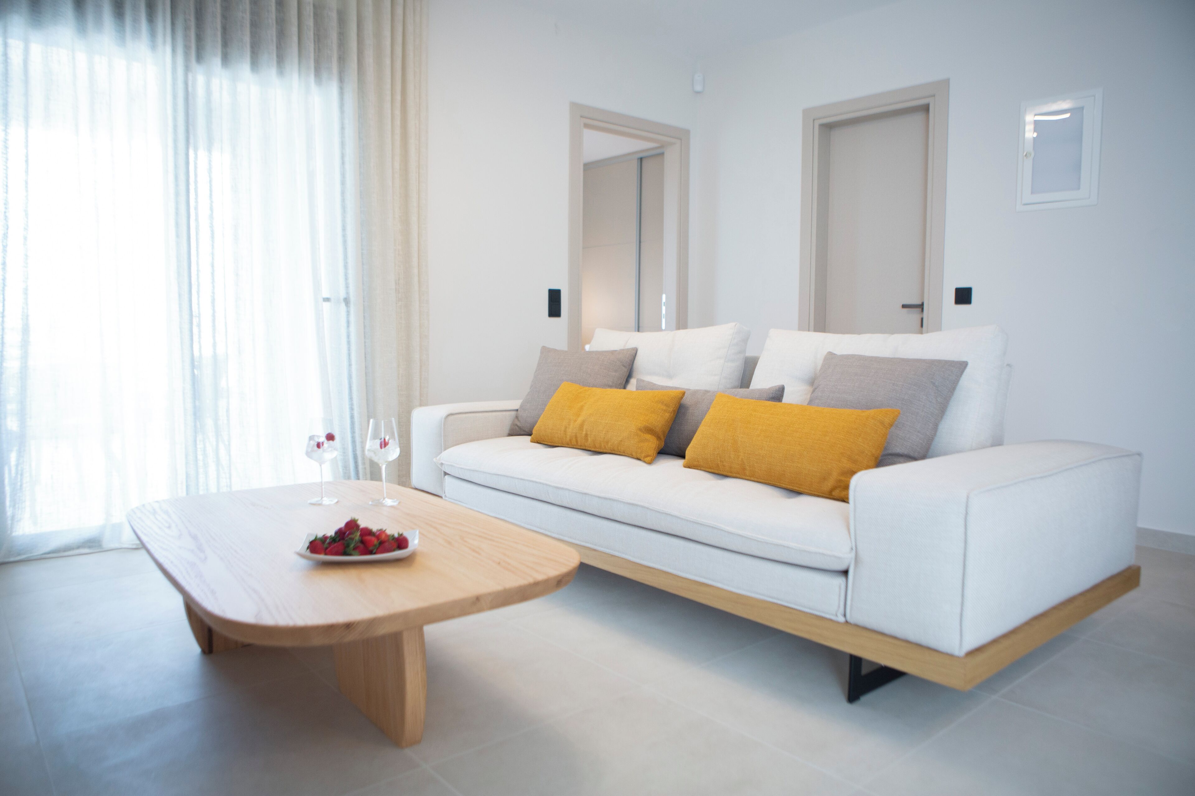 Un salon moderne avec un canapé blanc, des coussins jaunes, une table basse en bois sur laquelle est posé un panier de fruits, et une grande fenêtre ornée de rideaux légers.