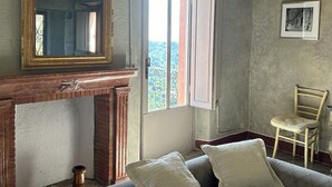Een kamer met een bank, een open haard, een spiegel, een raam met gordijnen, en een ingelijst schilderij aan de muur.