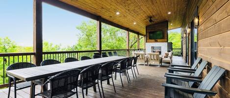 A covered outdoor dining area with a long table and chairs, a grill, and a TV mounted on the wall.
