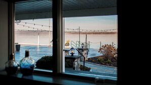 Vista desde el interior de un edificio hacia una masa de agua, con luces colgantes, un banco de piedra y plantas en macetas.