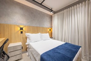 Habitación de hotel con cama, escritorio y silla. Cuenta con dos lámparas de pared y un ventanal con cortinas.