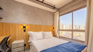 A hotel room with a bed, a desk, a chair, a nightstand, and a view of the city through large windows. - NOV - NEXT ONE Vila Nova Conceição (São Paulo)