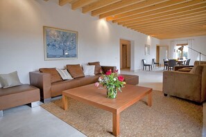 A modern living room with a wooden ceiling, a sofa set, a coffee table with a vase of flowers, and a dining area in the background.
