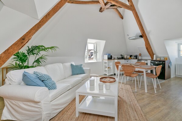 A loft-style living space with a white sofa, dining table, and chairs, a kitchen area, and a large window.