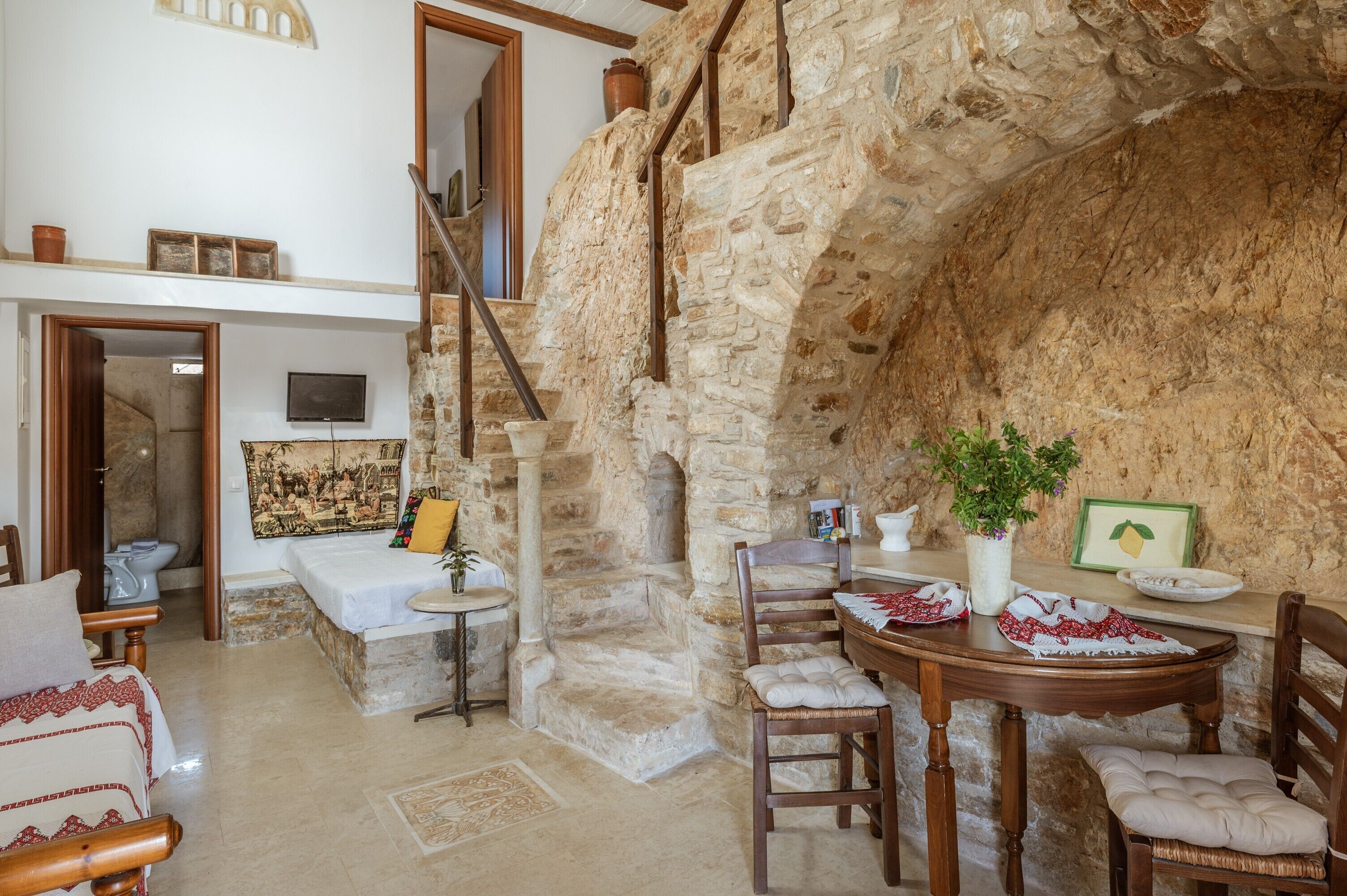 A rustic room with a stone wall, wooden staircase, a bed, a dining table with chairs, and a small table with a plant.