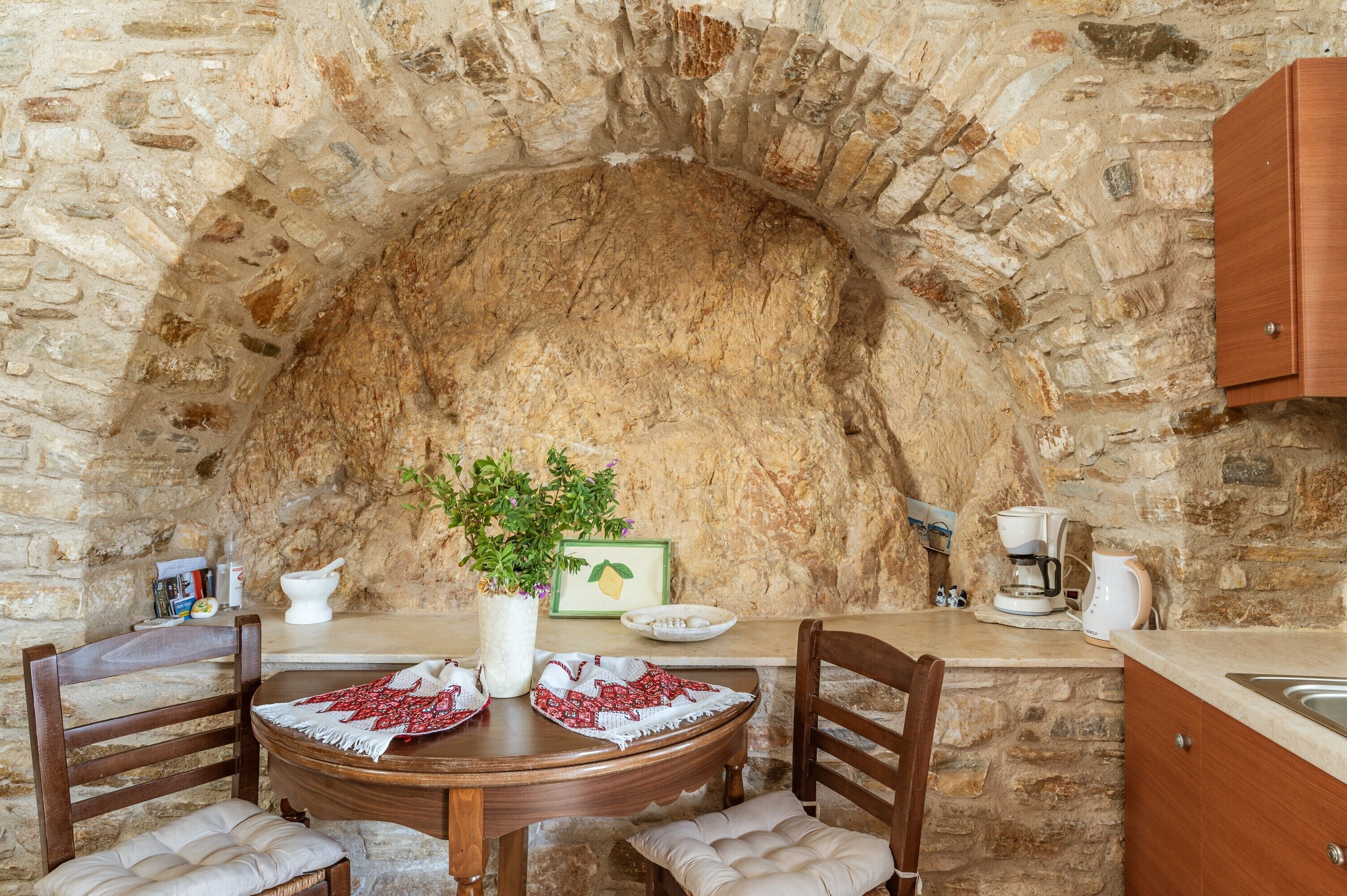 A stone-walled room with a round table, chairs, a coffee machine, and a cabinet.