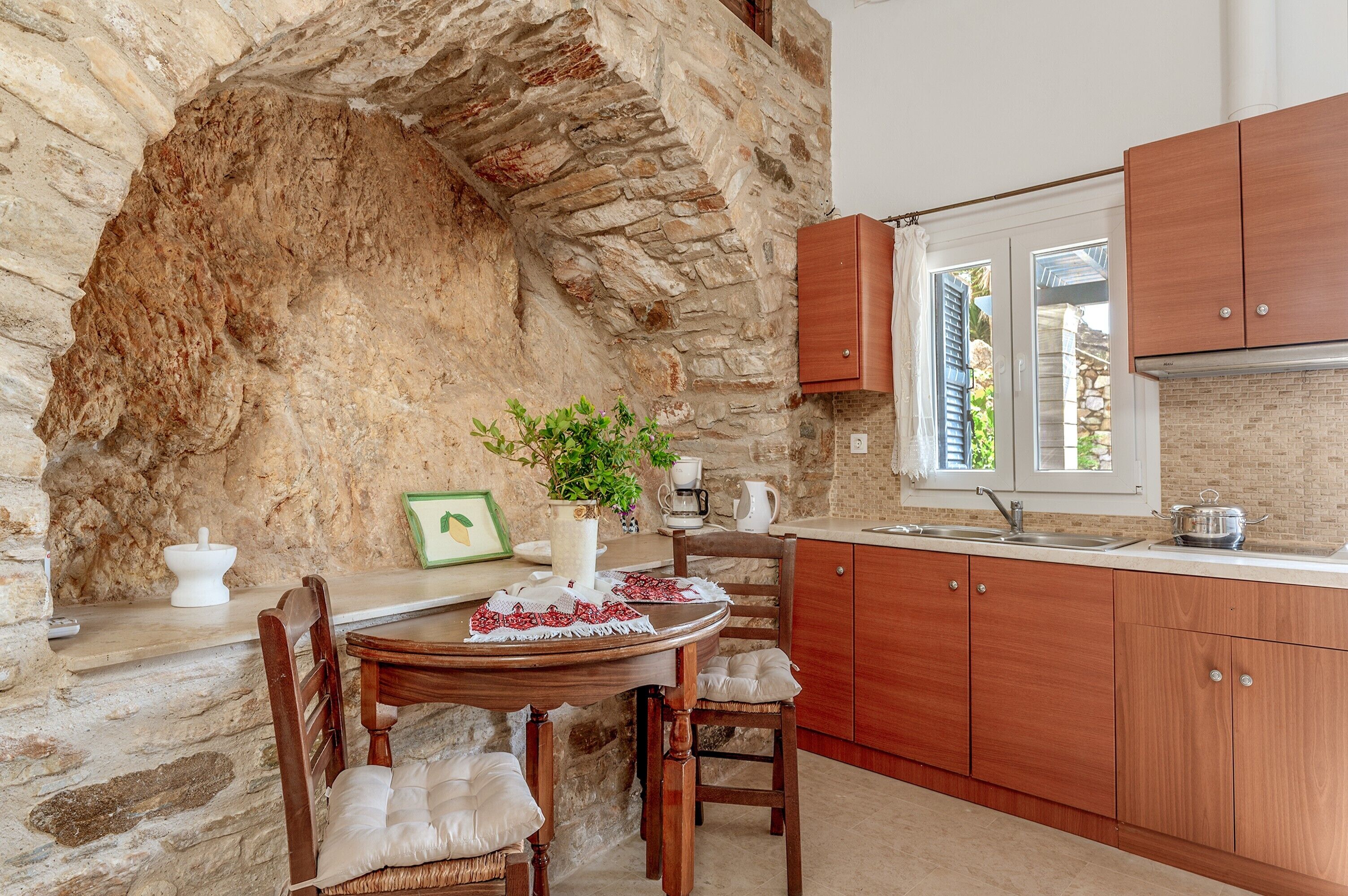 A stone-walled kitchen with wooden cabinets, a dining table with chairs, and a window with shutters.