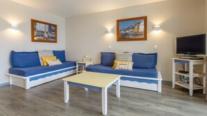 A living room with two blue sofas, a small white table, a TV on a stand, and two wall-mounted lights. - Apartment 'Sea View on the Beach' with Sea View, Shared Pool and Wi-Fi (Douarnenez)