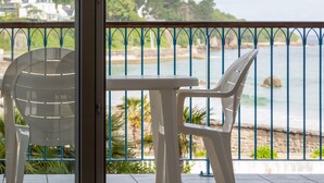 A balcony with a table and two chairs, overlooking a coastal area with a stone wall and greenery. - Apartment 'Sea View on the Beach' with Sea View, Shared Pool and Wi-Fi (Douarnenez)
