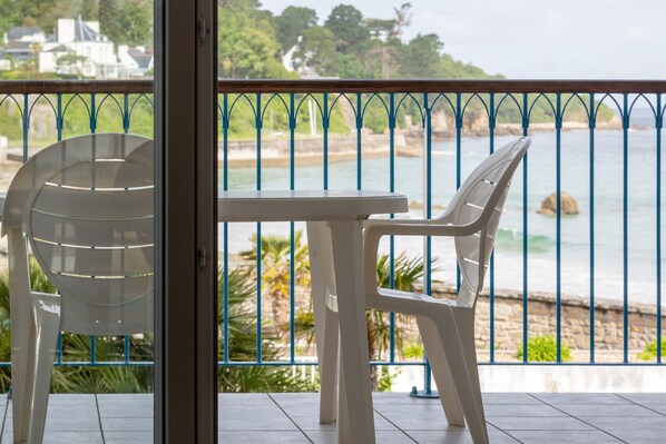 A balcony with a table and two chairs, overlooking a coastal area with a stone wall and greenery.