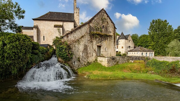 A historic stone building with a waterfall, surrounded by greenery and a clear sky.
