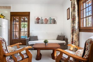 A room with a wooden table, two chairs, a sofa, a painting of vases, and a window with curtains.