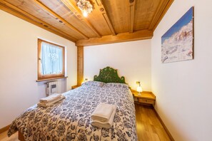 Une chambre avec un plafond en bois, un lit recouvert d’une couvre-lit à motifs, une fenêtre avec un rideau, une table de chevet avec une lampe et un tableau accroché au mur.