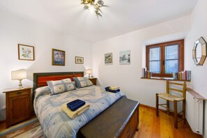 Un dormitorio con cama de madera, una mesita de noche con lámpara, una silla y una ventana con libros en un estante.