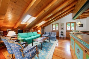 Un loft spacieux avec un plafond haut, des poutres en bois, un canapé vert, des fauteuils bleus, une table basse et un coin cuisine.