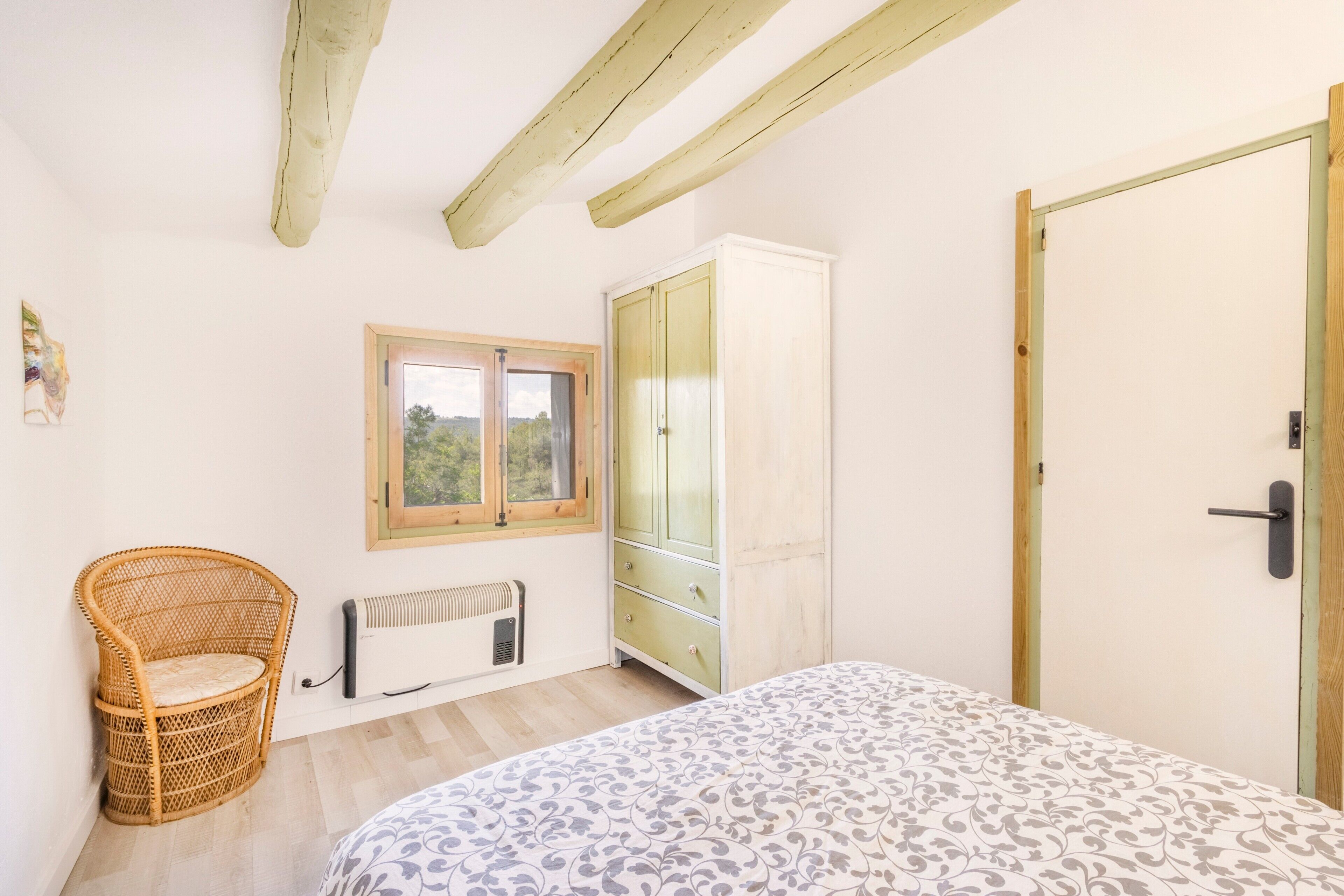 Un dormitorio con cama, una silla de mimbre, un armario, un radiador y una ventana.