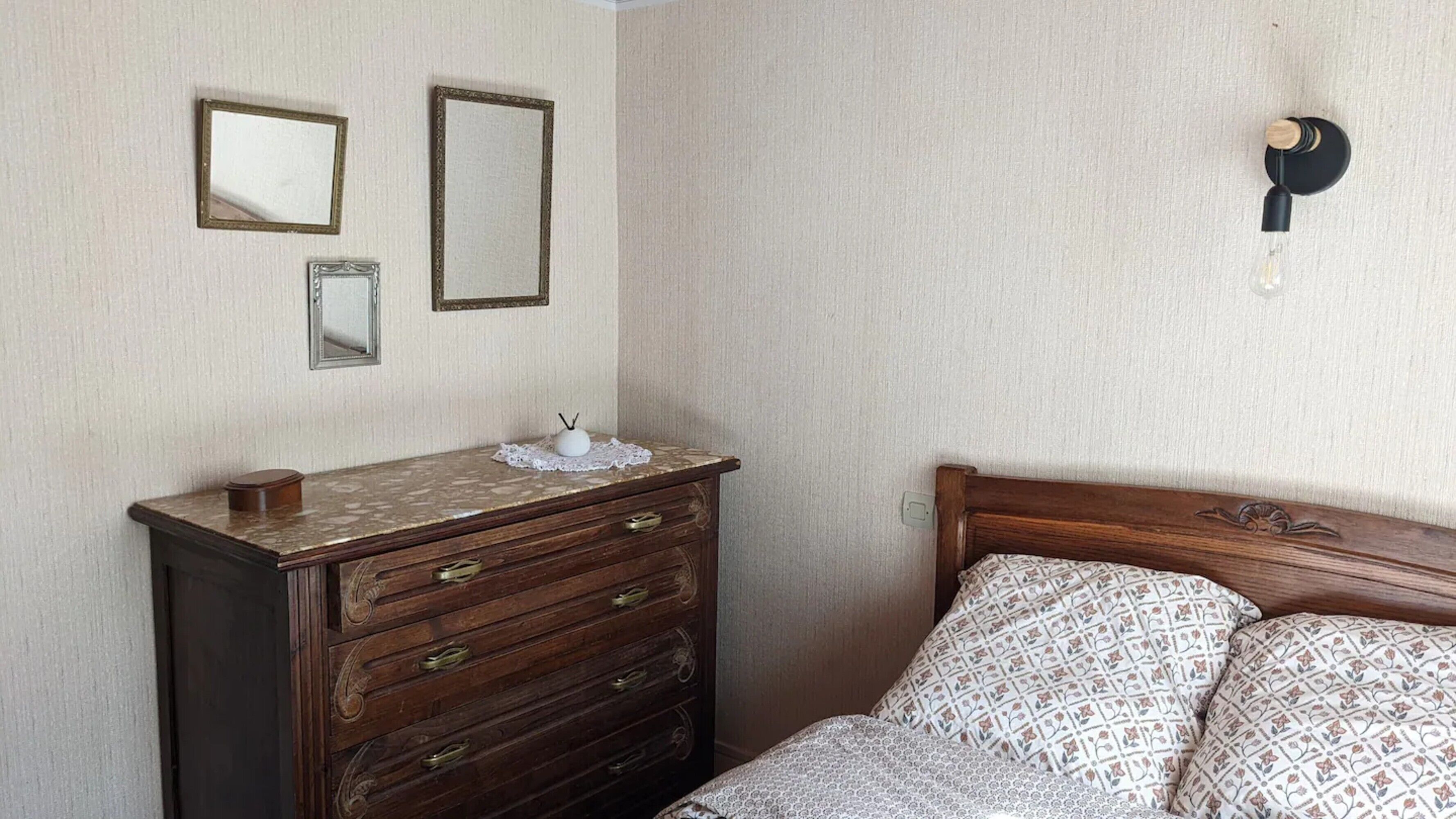A bedroom with a wooden dresser, two wall mirrors, a bedside table with a lamp, and a patterned bedspread.