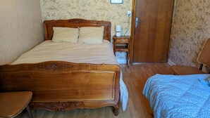 Uma cama de madeira com cabeceira, uma mesa de cabeceira com abajur, uma cadeira e uma porta.