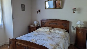 Une chambre avec un lit en bois, une literie à motifs floraux, une table de chevet avec une lampe, une commode, un miroir et un luminaire fixé au mur.
