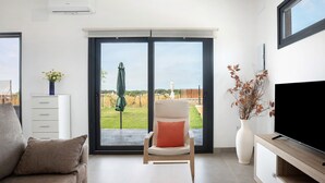 A modern living room with a sofa, a chair with a cushion, a flat-screen TV, a white cabinet, a vase with dried plants, and a sliding glass door leading to a garden. - Holiday Home 'Casa Valle' with Private Pool, Wi-Fi and Air Conditioning (Conil de la Frontera)
