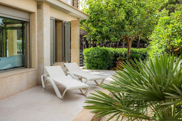 A patio with two white lounge chairs, a large window, and a well-maintained garden with various plants.