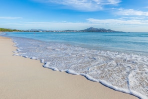 A beach with clear water, a mountain in the background, and a clear sky.