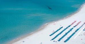 Vue aérienne d’une plage bordée de rangées de parasols bleus et de sable blanc.