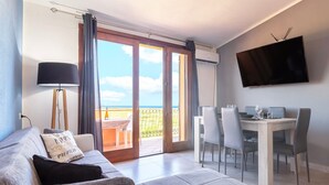 A modern living room with a sofa, a dining table with chairs, a TV mounted on the wall, and a sliding glass door leading to an outdoor area. - Holiday Home 'Calypso Sea View Holiday House 2' with Sea View, Wi-Fi and Air Conditioning (Badesi)