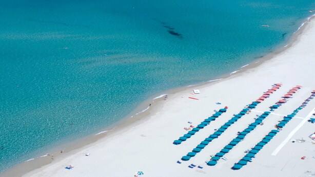An aerial view of a beach with clear turquoise water, rows of blue umbrellas, and a sandy shoreline.