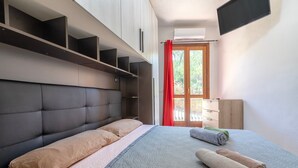 A modern bedroom with a large bed, built-in shelves, a flat-screen TV, and a sliding glass door to the outside.