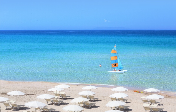 Una playa con agua azul cristalina, un velero y filas de sombrillas y tumbonas.