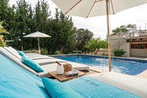 Un área junto a la piscina con sillones de descanso, una mesa de madera con un sombrero y un libro, y una sombrilla grande.