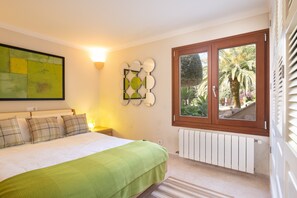 Une chambre avec un lit, une lampe fixée au mur, une fenêtre donnant sur des palmiers et un radiateur.