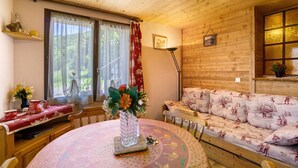 Interior acogedor de una cabaña con techo de madera, una mesa redonda de comedor con mantel floreado, un sofá con cojines estampados y un jarrón con flores sobre la mesa.