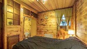 Una habitación de cabaña de madera con una cama, un ventanal con cortinas, una silla y una mesita de noche con una lámpara.