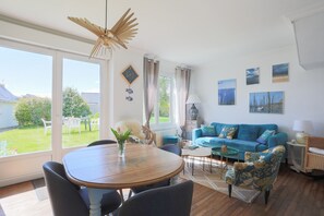 Een woonkamer met een blauwe bank, een houten eettafel en uitzicht op de tuin door grote ramen.