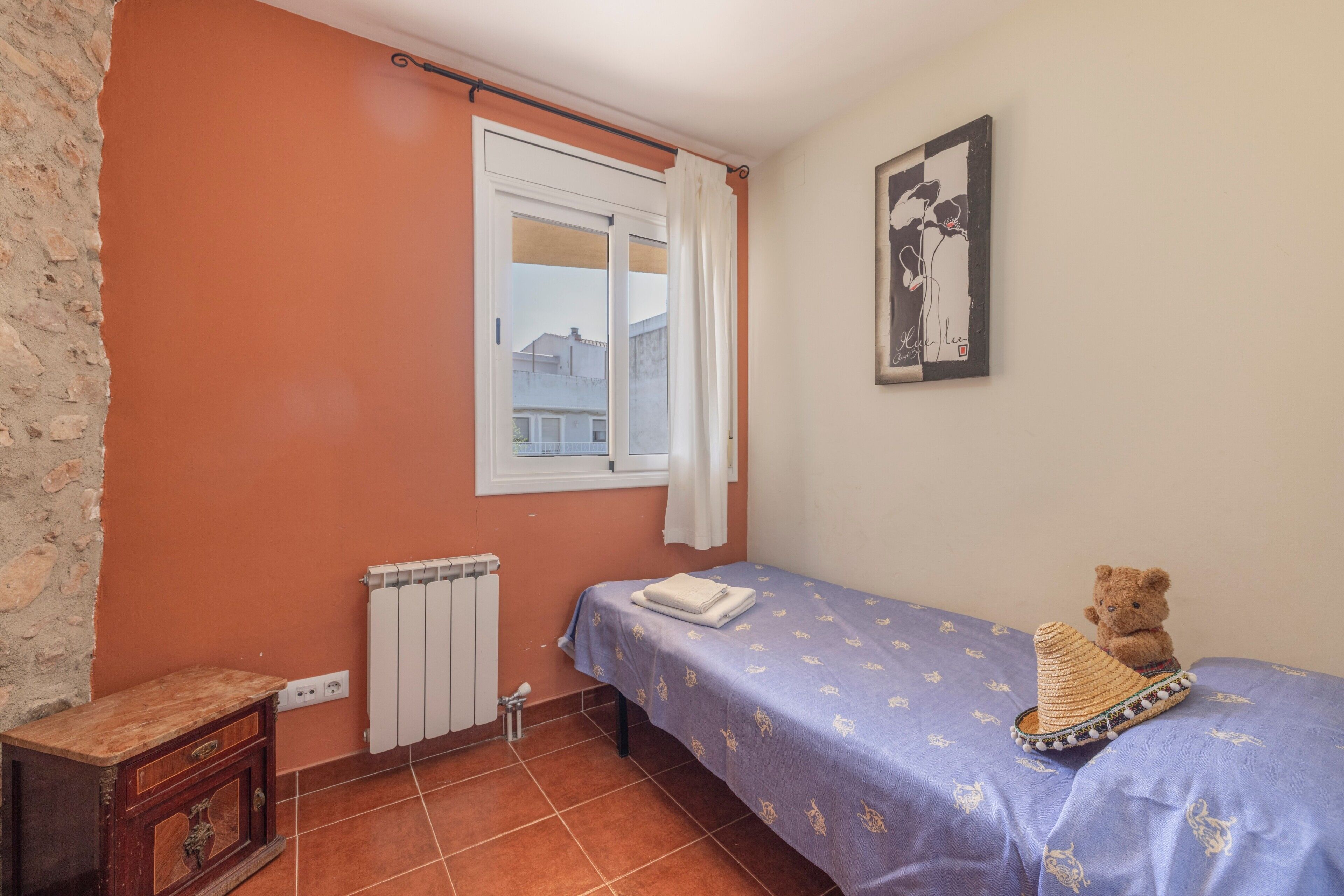 Ein Schlafzimmer mit einem Bett, einem Teddybären, einem Heizkörper, einem Fenster mit Vorhängen und einem Wandbild.