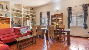 Una sala de estar con un sofá rojo, una mesa de centro de madera, sillas de mimbre, una mesa de comedor y estanterías con diversos objetos.