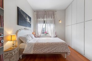A bedroom with a bed, a nightstand, a lamp, a window with floral curtains, and a painting on the wall. - Holiday Home 'Romantic Apartment Metro Flaminio' with Wi-Fi and Air Conditioning (Roma)