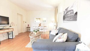 Una sala de estar con un sofá gris, un televisor sobre un mueble bajo, una mesa de comedor con sillas y un cuadro del Coliseo colgado en la pared.